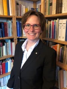 Eine Person mit Brille, schwarzem Blazer und weißem Hemd steht lächelnd vor den mit Büchern gefüllten Bücherregalen auf dem Campus Klarenthal.