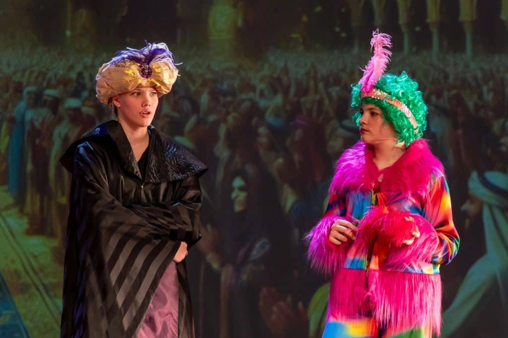 Zwei Personen auf der Bühne des Campus Klarenthal tragen bunte, aufwendige Kostüme und Kopfbedeckungen. Die eine trägt ein schwarzes Gewand mit einem goldenen Turban, die andere glänzt in Pink und Grün mit einem Federkopfschmuck. Hinter ihnen wird eine Massenszene projiziert.
